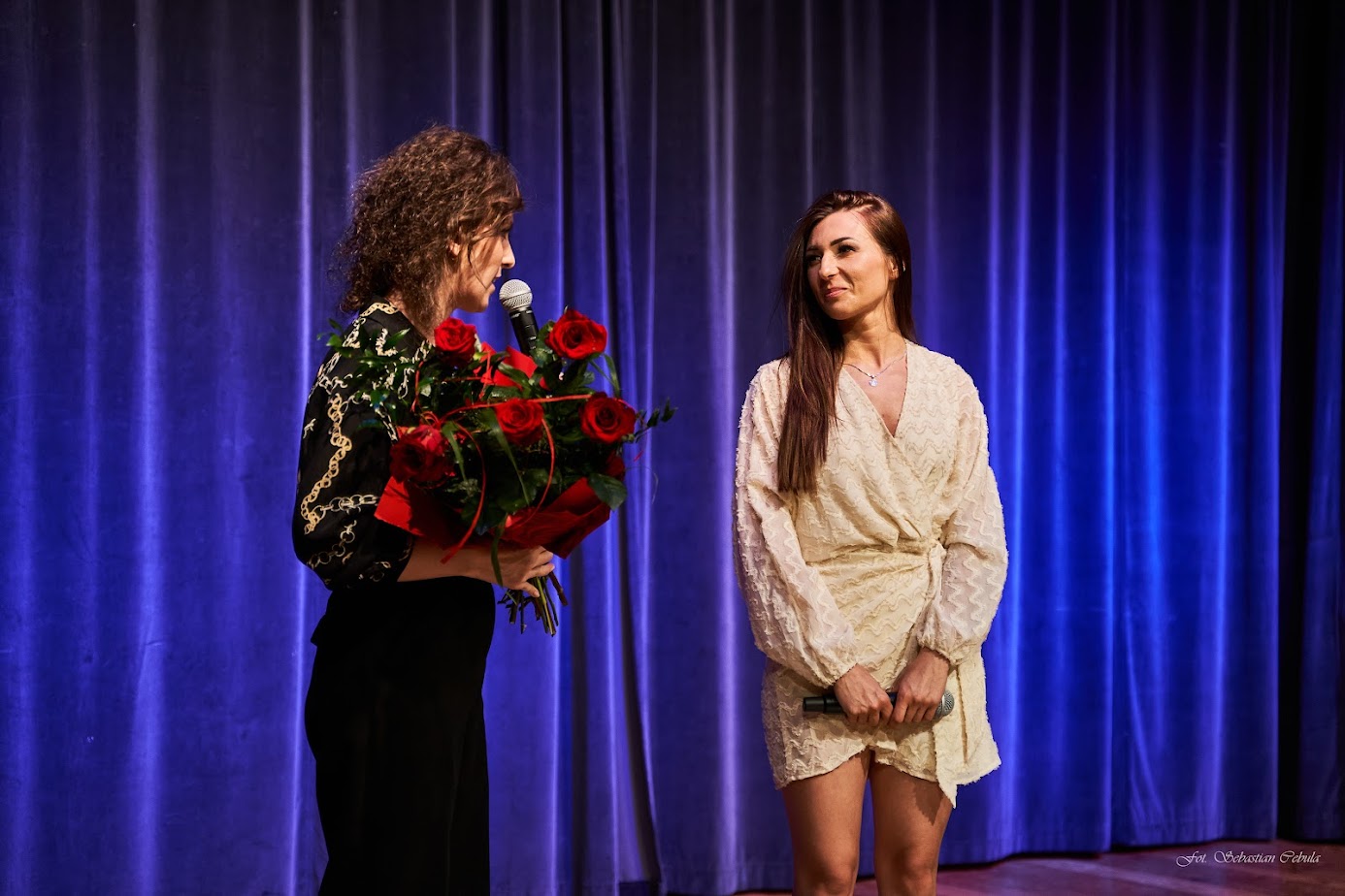 na scenie stoją dwie kobiety jedna trzyma bukiet czerwonych róż
